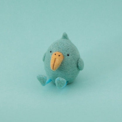 A small handmade felt bird toy in a light blue-green color with a contrasting orange beak, sitting against a solid light blue-green background.